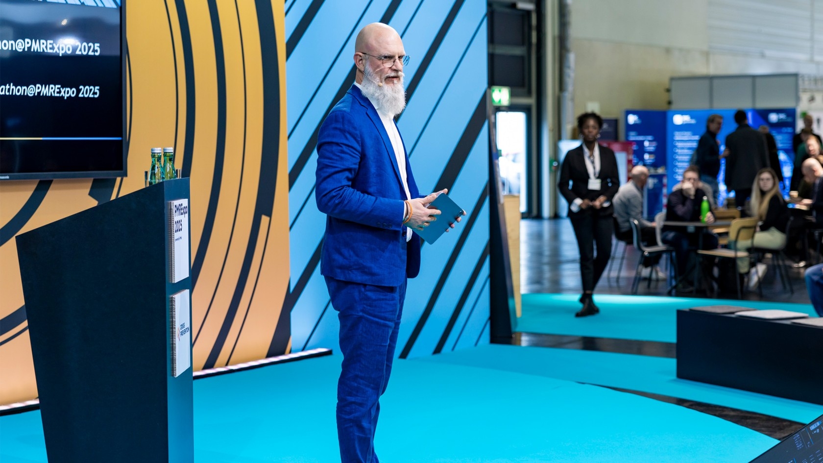 Ein bärtiger Mann in einem blauen Anzug steht auf der Bühne mit einem Tablet in der Hand und präsentiert vor einem Publikum beim PMRExpo 2025, das im Hintergrund sichtbar ist.