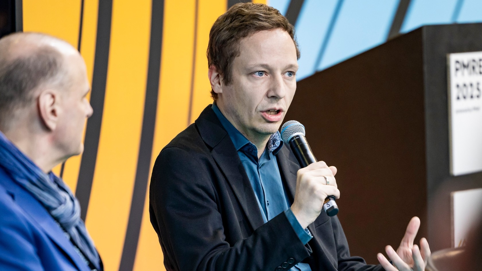 Ein Redner mit einem Mikrofon erörtert ein Thema während einer Podiumsdiskussion, während ein anderer Teilnehmer aufmerksam in der professionellen Atmosphäre der Konferenz zuhört.