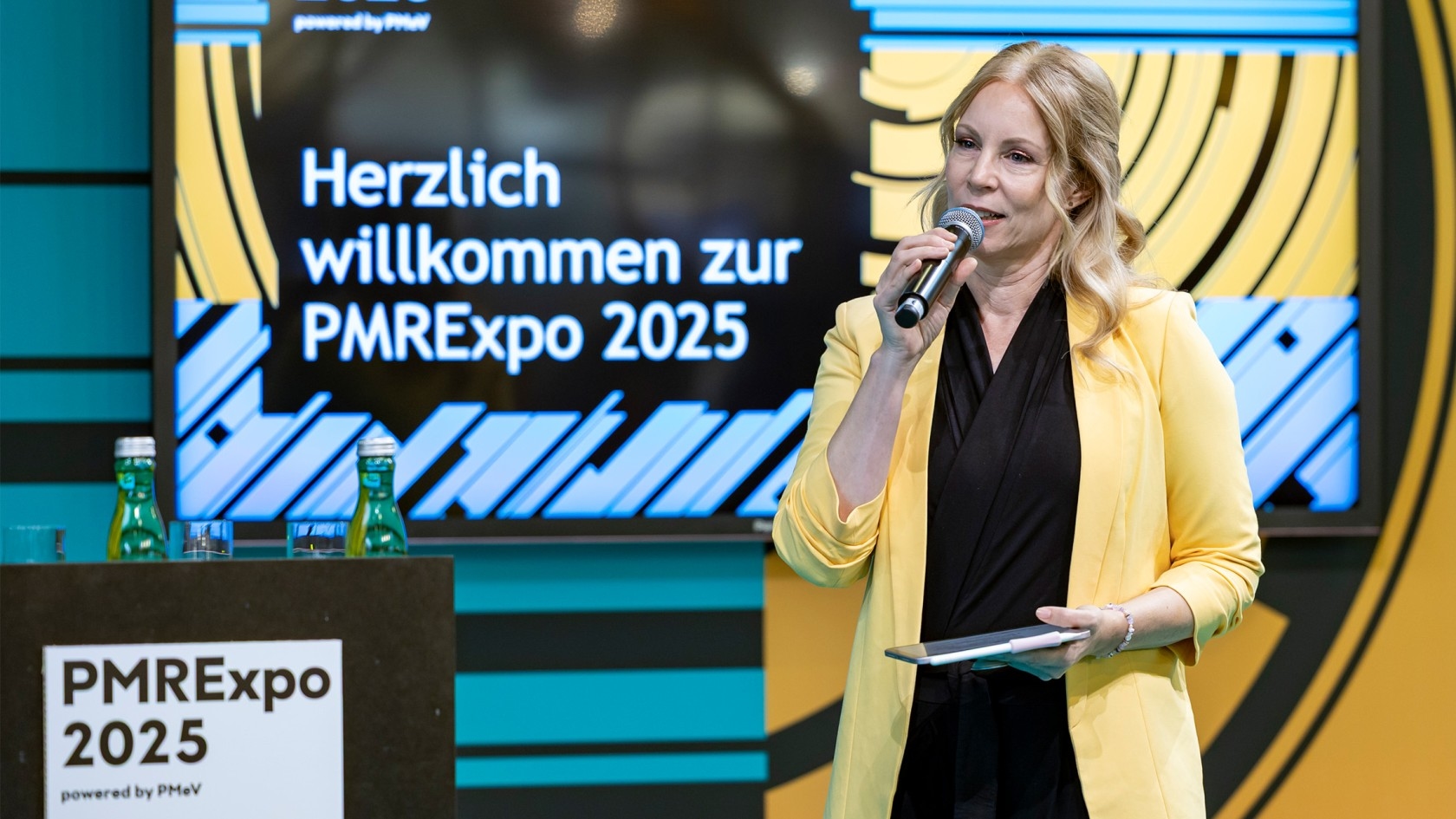 Eine Frau in einem gelben Blazer spricht auf der Bühne in ein Mikrofon, während im Hintergrund "PMRexpo 2025" sowie eine Willkommensnachricht zu sehen sind.