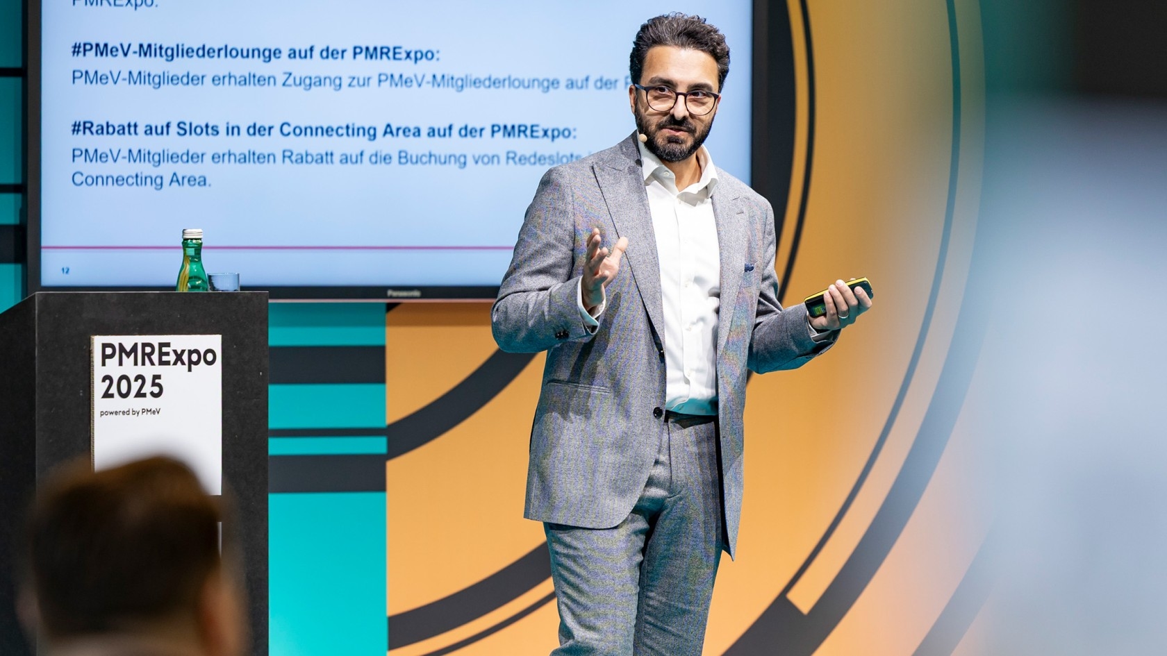 Ein Referent in einem grauen Anzug gestikuliert lebhaft während seiner Rede auf der Fachkonferenz, wobei eine Folie zu PMRExpo 2025 im Hintergrund zu sehen ist.