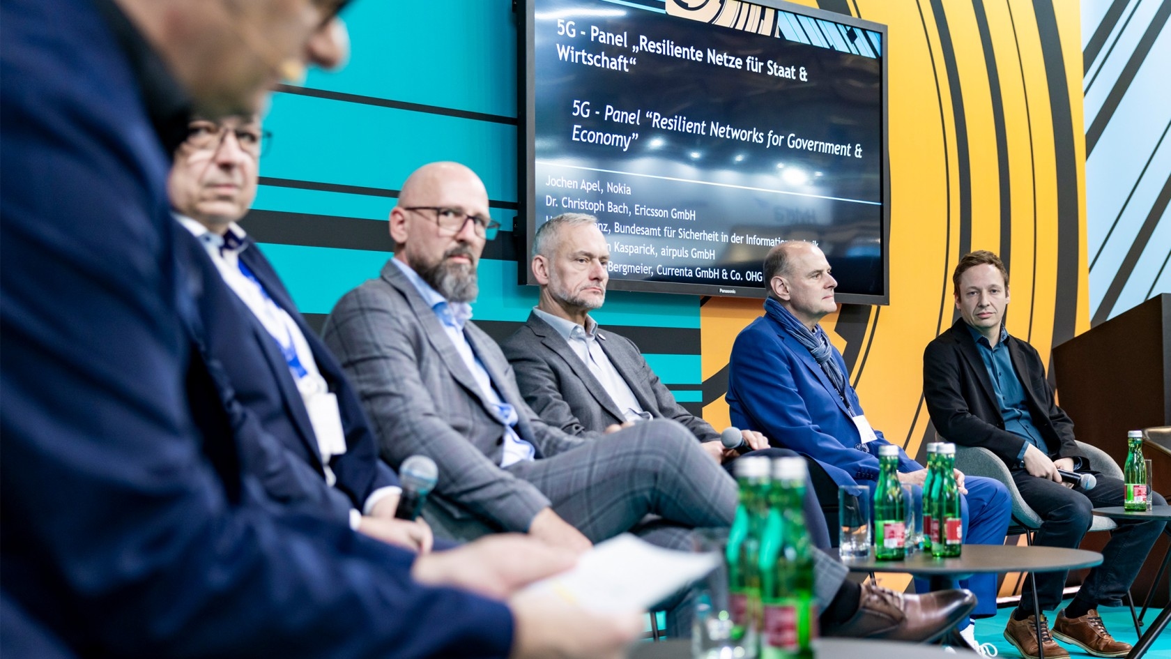 Ein lebhafter Paneldiskussion über resiliente Netzwerke für Regierung und Wirtschaft, bei dem fünf Sprecherinnen und Sprecher anwesend sind, während auf den Tischen erfrischende Getränke bereitstehen und ein digitaler Präsentationsbildschirm die wichtigsten Punkte visuell unterstützt.