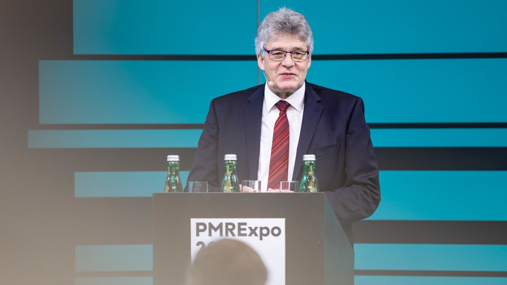 Ein Redner in einem Anzug präsentiert am Podium mit Wasserflaschen während der PMRexpo 2023 Veranstaltung vor einem eindrucksvollen blauen Hintergrund.