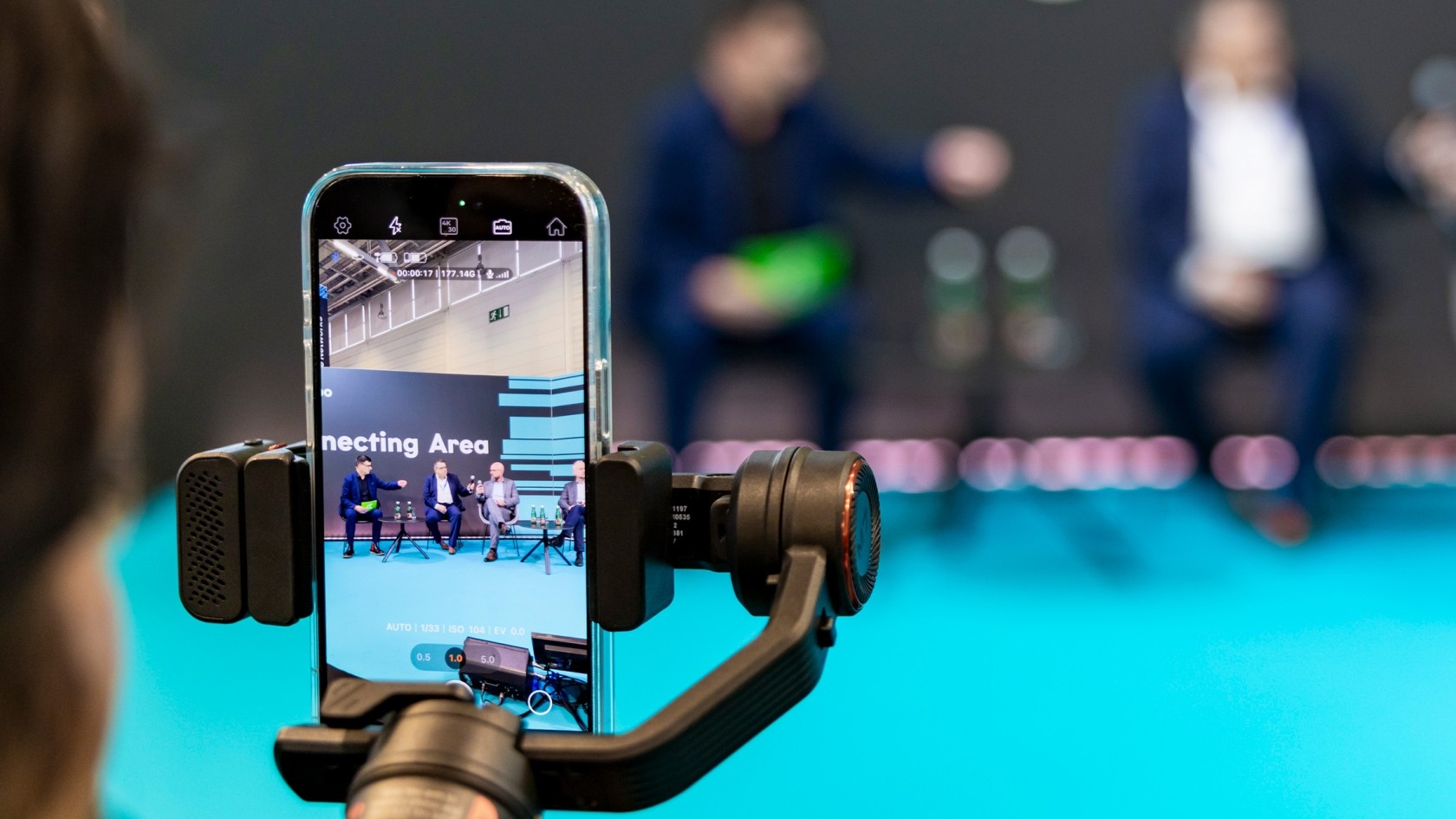 Ein Smartphone auf einem Stabilisator filmt eine Podiumsdiskussion mit Sprechern auf der Bühne, vor einem Hintergrund, der "Verbundene Regionen" zeigt.