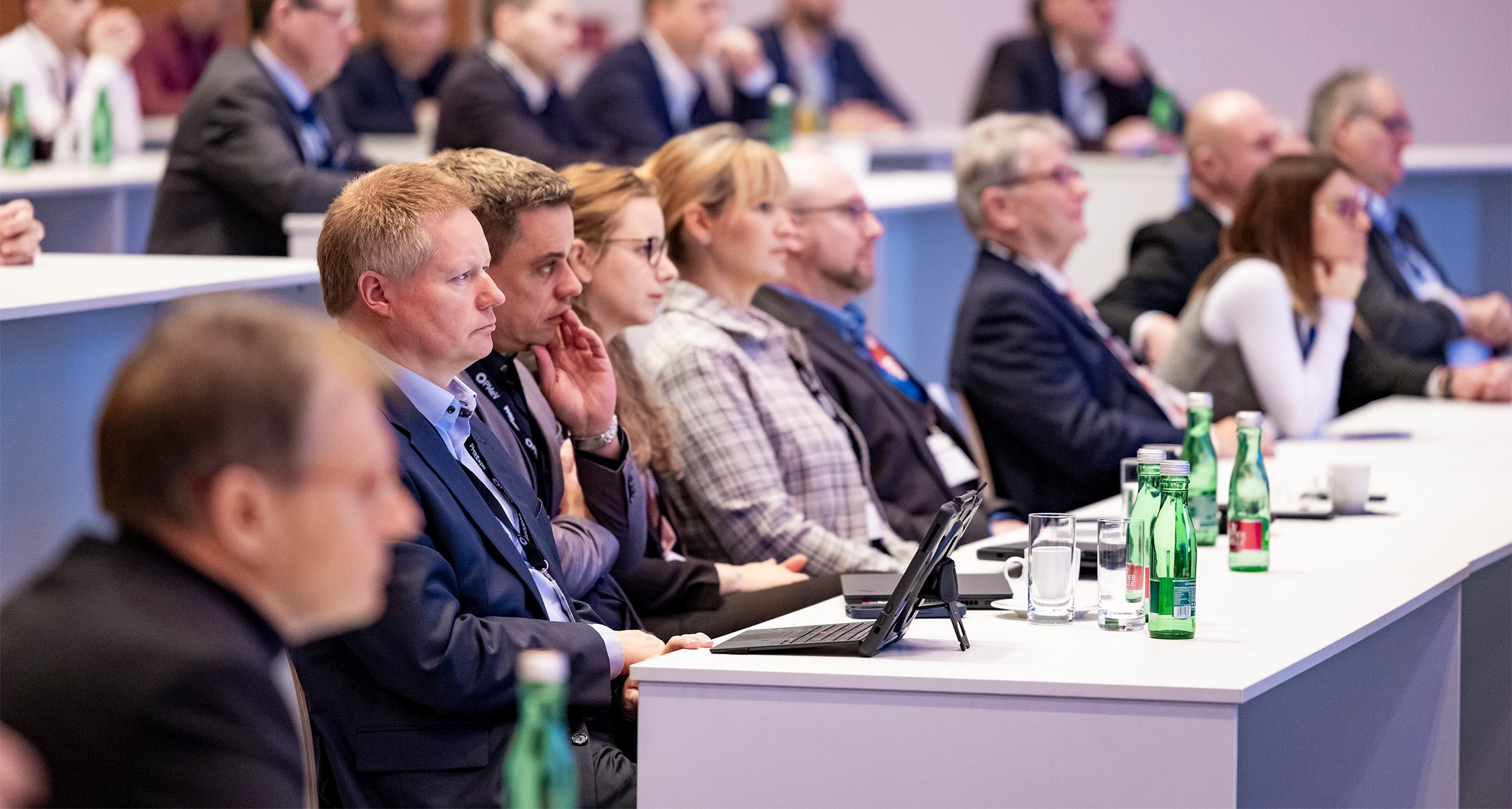 Die Teilnehmer sitzen aufmerksam in einem modernen Konferenzraum und blicken auf den Redner, während auf den Tischen Wasserflaschen und Gläser bereitstehen.