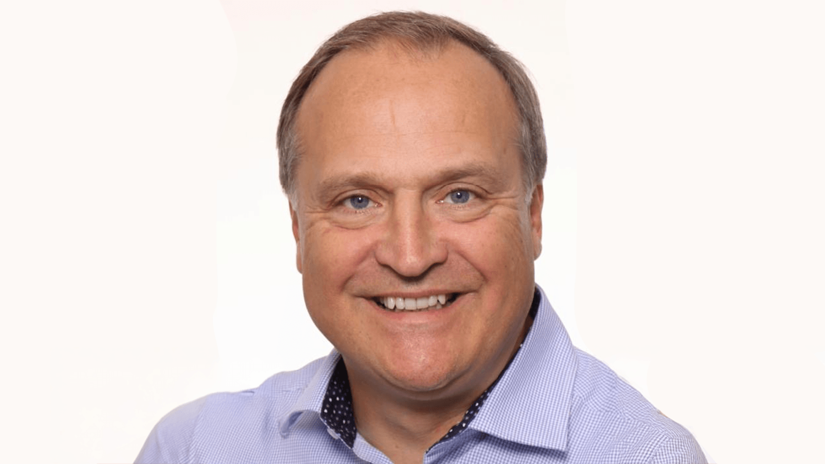 Ein lächelnder Mann mit kurzem Haar trägt ein hellblaues Hemd und steht vor einem schlichten weißen Hintergrund, während er bereit ist, innovative Kommunikationslösungen und Sicherheitskonzepte auf der PMRExpo zu präsentieren.