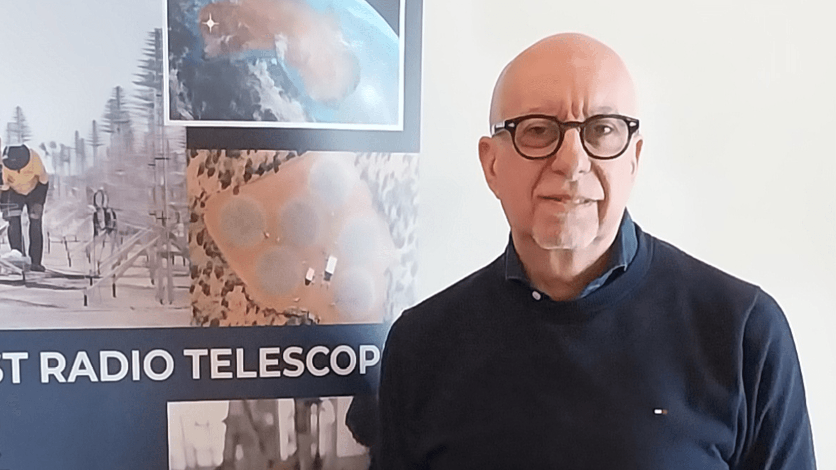 Ein Mann mit Brille steht vor einem Plakat, das verschiedene Antennenmodelle von Sirio Antenne zeigt, einem italienischen Hersteller, der innovative Technologien für das größte Radioteleskop der Welt präsentiert.