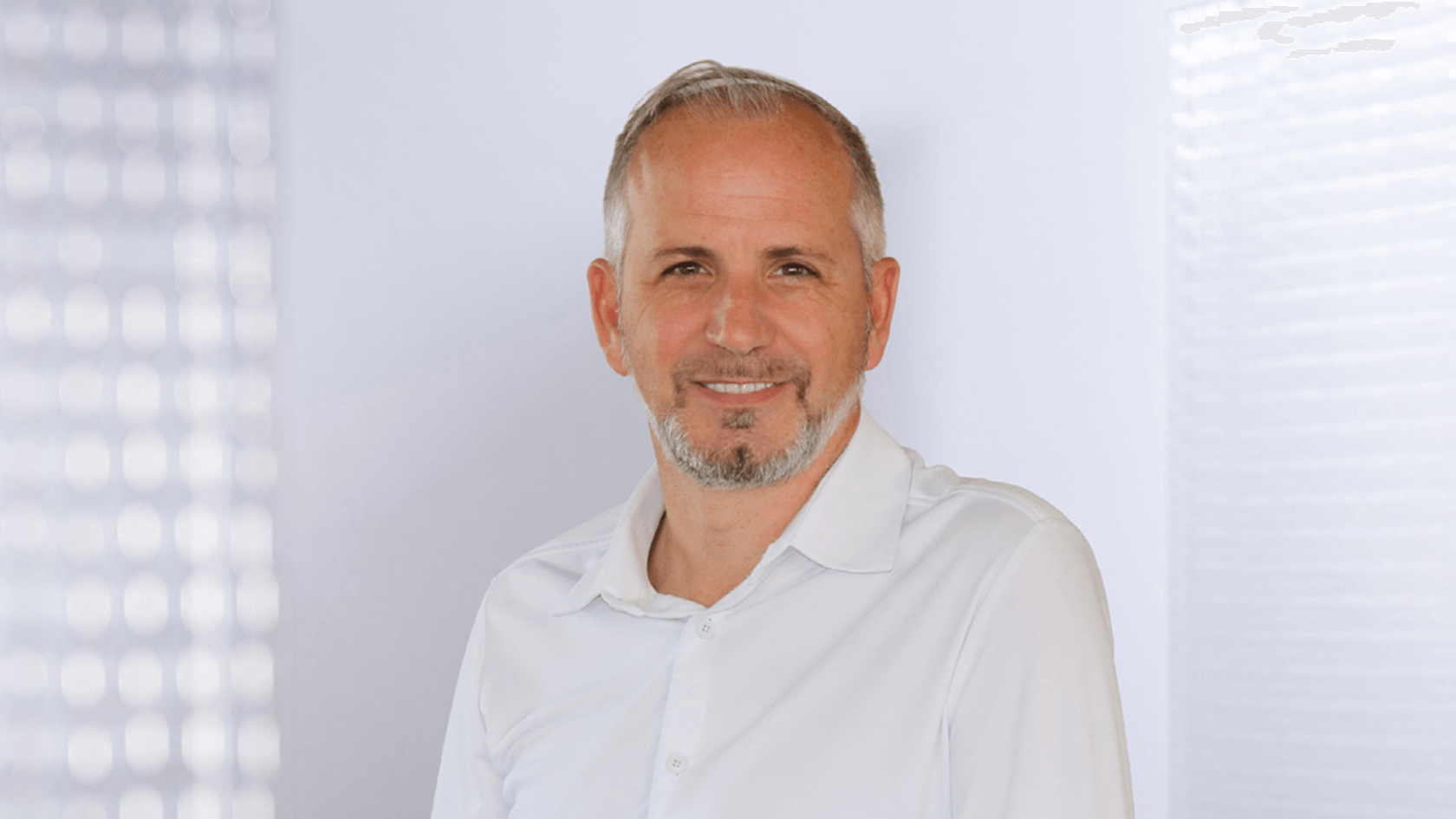 Ein lächelnder Javier Esparza, Verkaufsleiter für Europa bei RugGear, mit kurzem grauen Haar und einem Bart, steht in einem hellen Hemd vor einem sanften, neutralen Hintergrund während der PMRExpo, wo er die neuesten Entwicklungen in der kritischen Kommunikation präsentiert und partnerschaftliche Verbindungen knüpft.