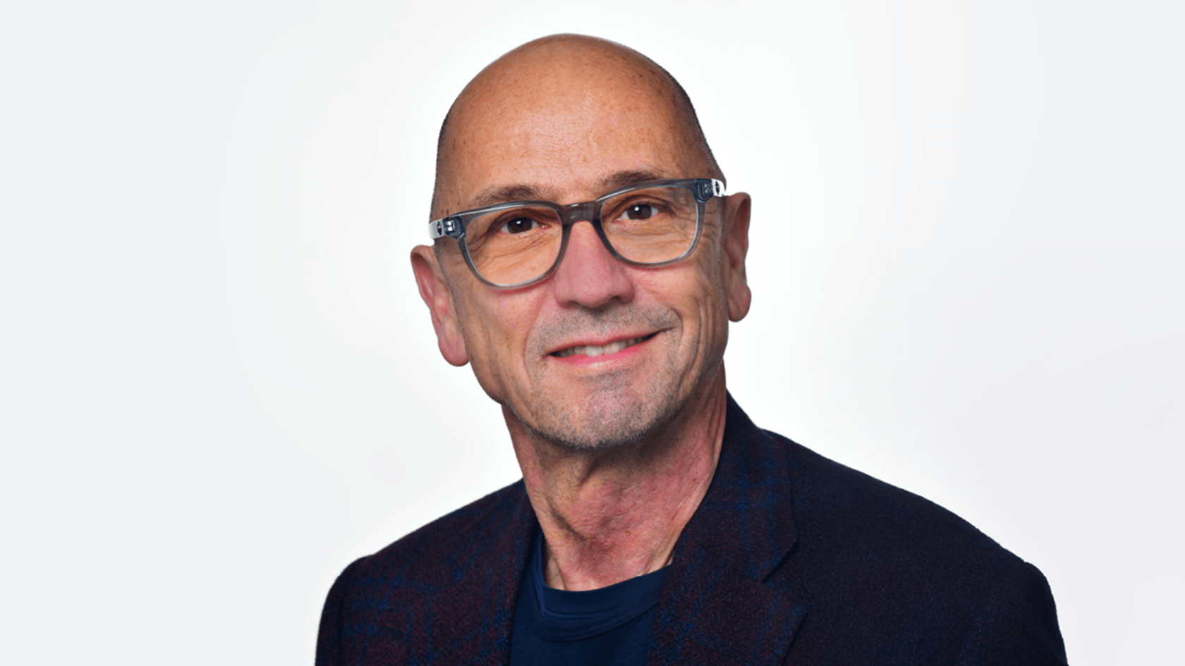 Ein lächelnder Mann mit einer Glatze und Brille, gekleidet in ein dunkles Sakko über einem marineblauen Hemd, präsentiert sich vor einem hellen Hintergrund, während er die bedeutenden Themen der PMRExpo reflektiert.
