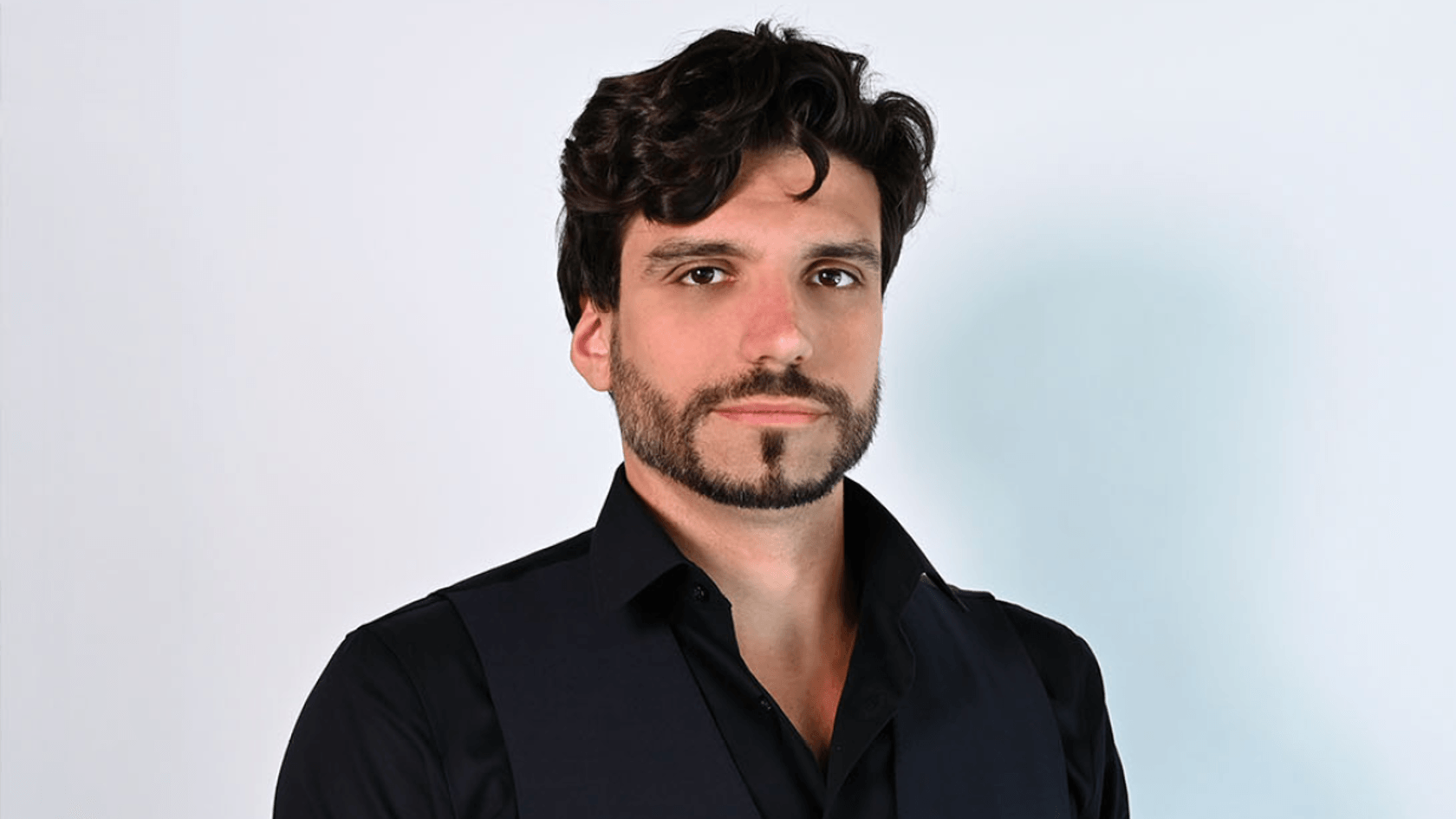 Ein Mann mit lockigem, dunklem Haar und einem Bart, der in einem schwarzen Hemd und Weste gekleidet ist, steht strahlend vor einem hellen Hintergrund und symbolisiert die Innovationskraft, die auf der PMRExpo für die Branche von Bedeutung ist.
