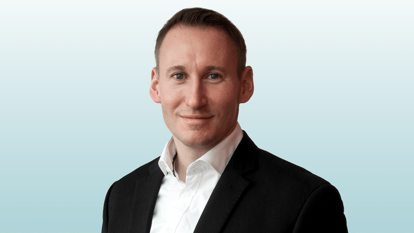 Ein lächelnder Mann in einem schwarzen Anzug und weißem Hemd posiert selbstbewusst vor einem hellen, sanften Hintergrund, während er die innovativen Lösungen seines Unternehmens auf der PMRExpo präsentiert.