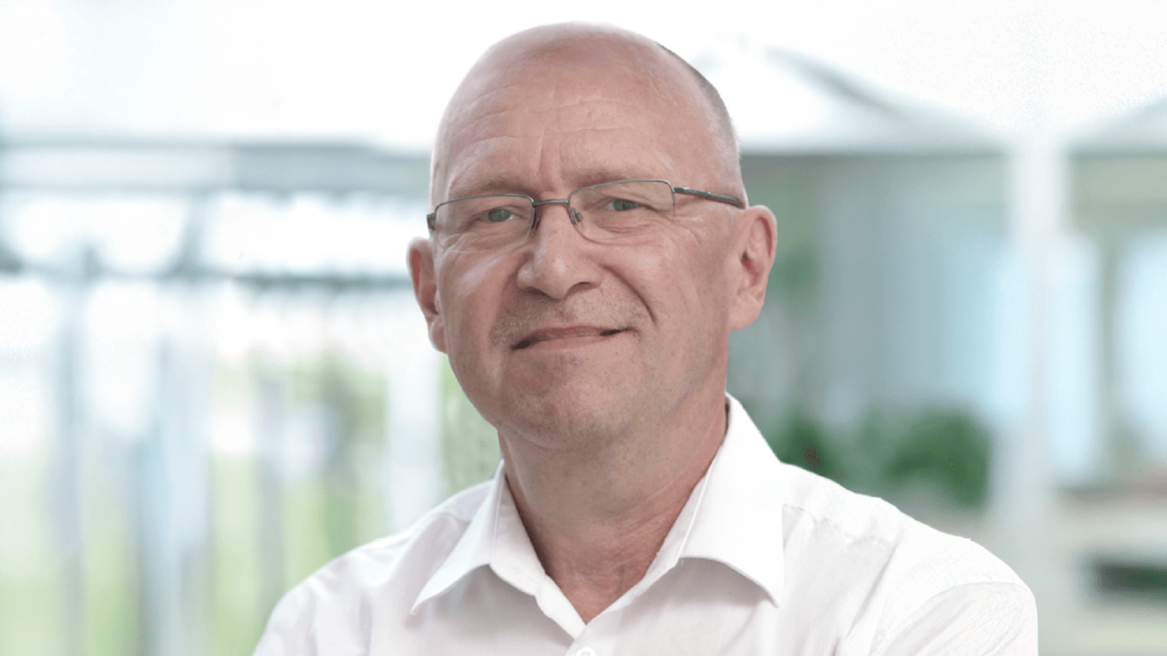 Ein mittelalter Mann mit Brille lächelt warmherzig in einem hellen, modernen Messeumfeld, während er ein weißes Hemd trägt und von üppigem Grün umgeben ist, was die lebendige Atmosphäre der PMRExpo unterstreicht.