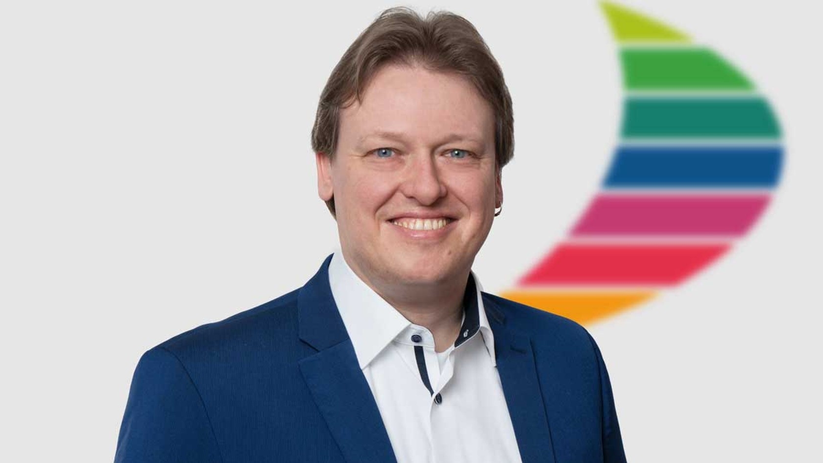 Ein lächelnder Jens Lenzing in einem blauen Anzug steht vor einem hellgrauen Hintergrund, während neben ihm teilweise das bunte Logo der PMRExpo zu sehen ist, die als zentraler Treffpunkt für BOS und Leitstellen dient.