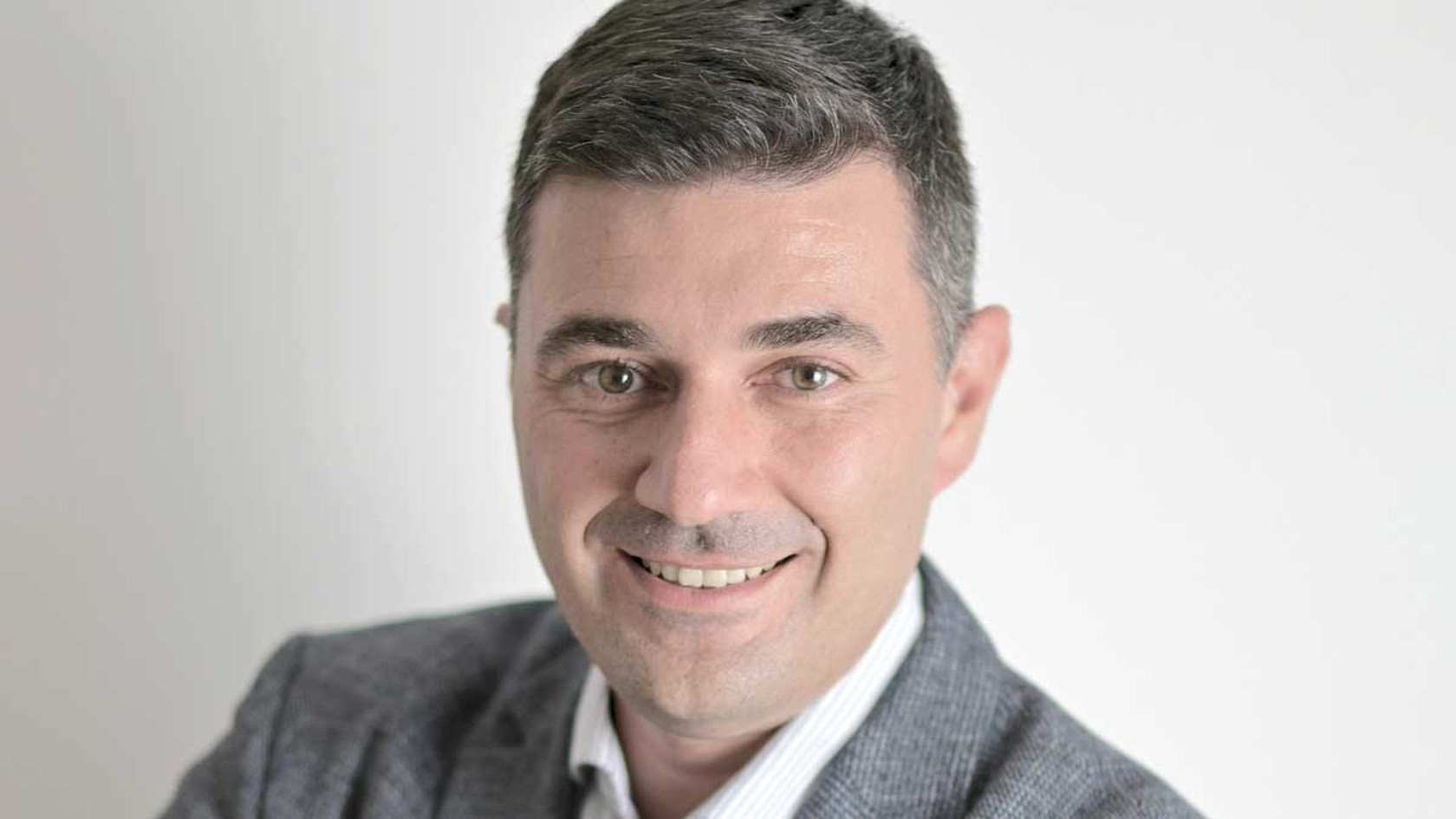 Ein lächelnder Mann mit kurzen dunklen Haaren und einem hellgrauen Blazer steht vor einem neutralen Hintergrund, während er stolz die Innovationen der Schnoor Industrieelektronik GmbH auf der PMRExpo präsentiert.