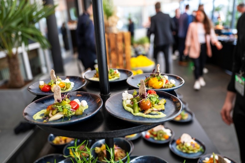 Catering Mehrere schwarze Teller mit kunstvoll angerichteten Vorspeisen auf einem mehrstöckigen Serviergestell in einem belebten Innenraum.