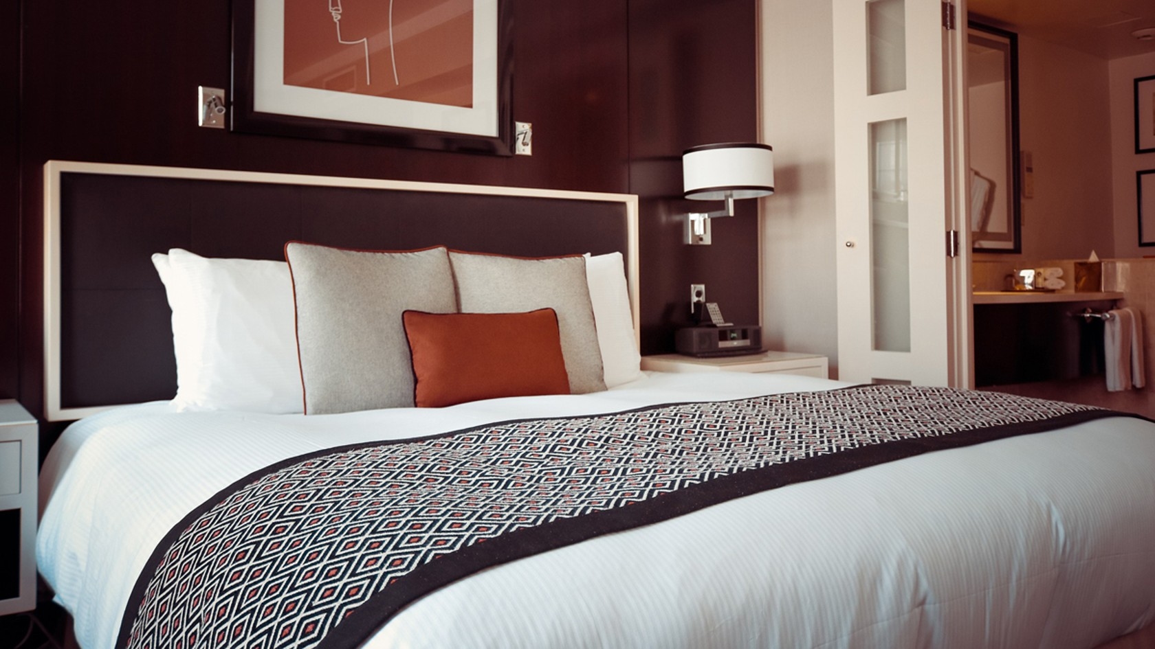 Hotelzimmer Doppelbett mit weißer Bettwäsche, grauen und orangefarbenen Kissen, geometrisch gemustertem Überwurf und dunklem Kopfteil in einem Hotelzimmer.