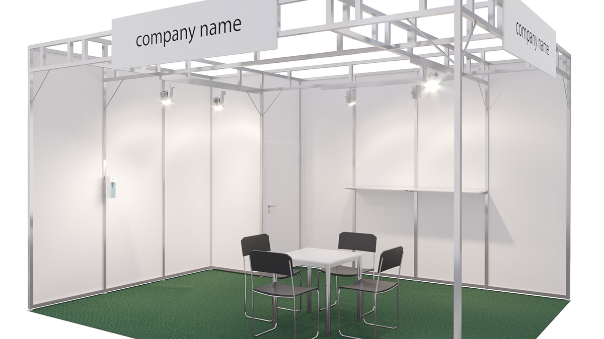 Ein schlichtes Ausstellungsstand-Design mit einem grünen Teppichboden, einem 1 m² großen Kabinett mit Garderobe, einem Möbelpaket bestehend aus einer Tischvitrine, zwei Barhockern und einem Prospektständer, sowie einer auffälligen Logo-Abbildung auf der Tischvitrine, beleuchtet von zwei Auslegestrahlern an der 250 cm hohen Hinterwand.