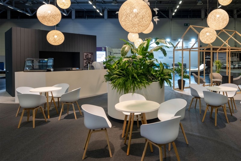 VIP Lounge Mehrere runde Tische mit weißen Stühlen umgeben einen großen Pflanzentopf in einem modernen Innenraum mit hängenden runden Lampen.